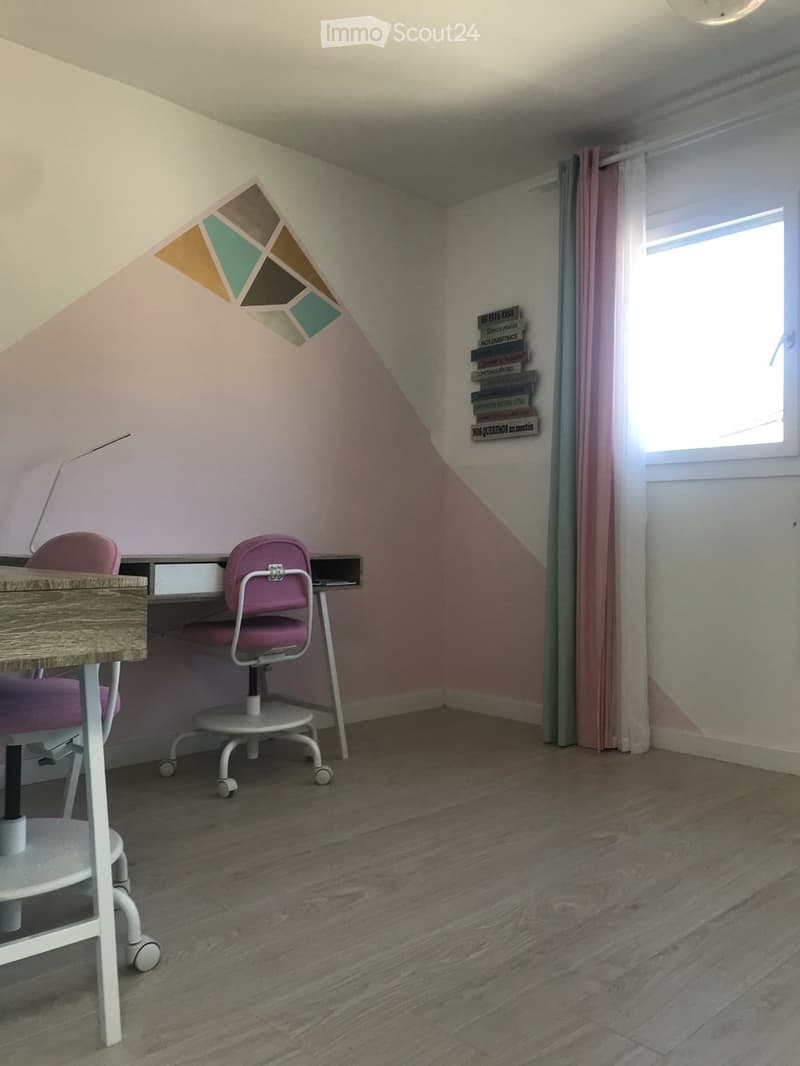 A small, cozy room with wooden flooring, a slanted ceiling, and a desk with two chairs. A window with a curtain is on the right side, and a stack of books is placed on the wall near the window.