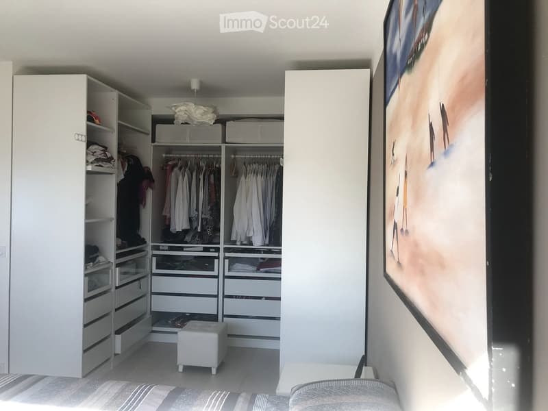Modern bedroom with a large wardrobe, neatly organized with hanging clothes and drawers, a small white stool, and a large picture frame on the wall.