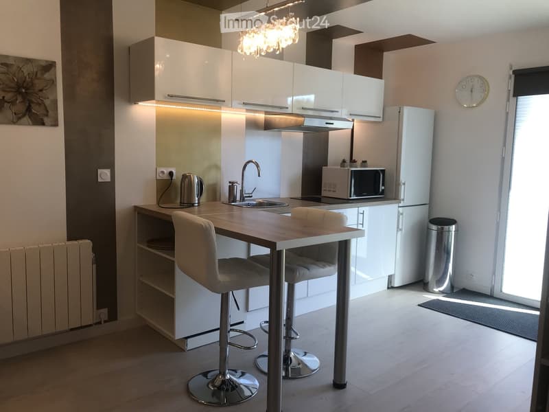 Modern kitchen with white cabinets, a central island with a wooden countertop, two bar stools, a sink, a microwave, and a refrigerator. The space is well-lit with pendant lighting and has a sleek, contemporary design.