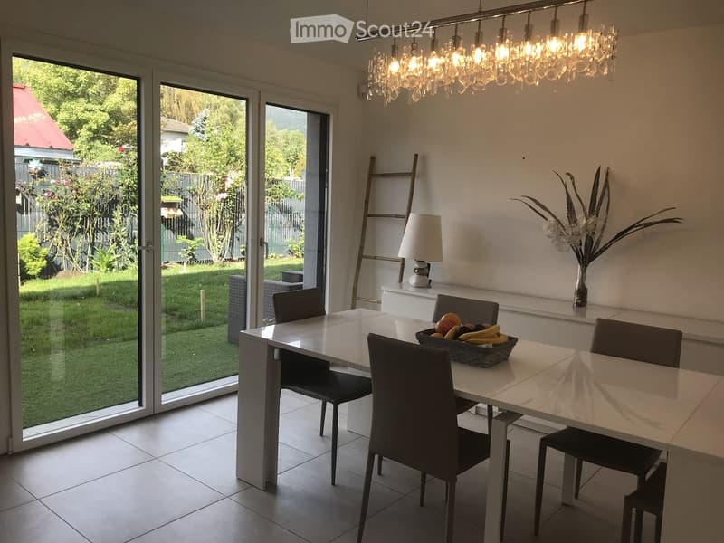 Modern kitchen with a white dining table, four chairs, a fruit basket, a table lamp, and a vase with flowers. Large glass doors open to a green garden.