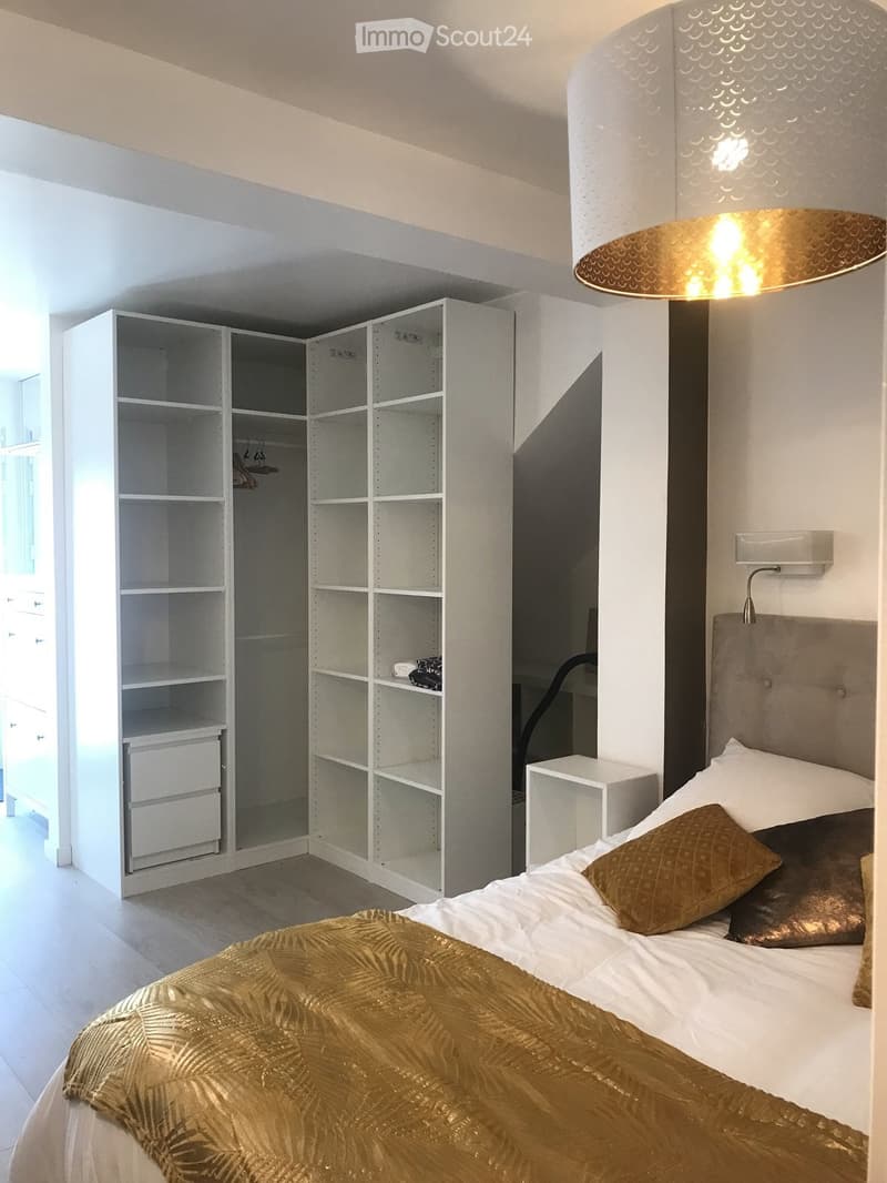 Modern bedroom with a large bed, gold throw, pillows, white wardrobe, shelves, and a hanging lamp.