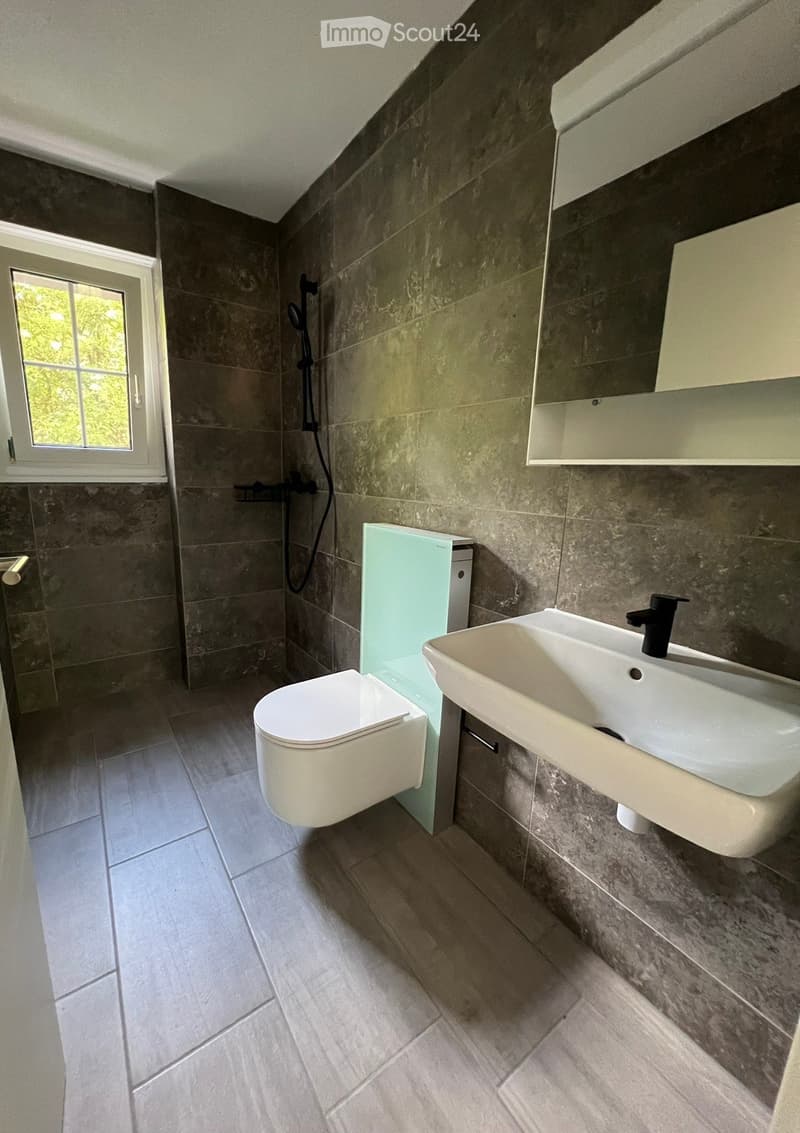Modern bathroom with a toilet, sink, mirror, and shower. Features grey tiles and a window with a view of greenery.