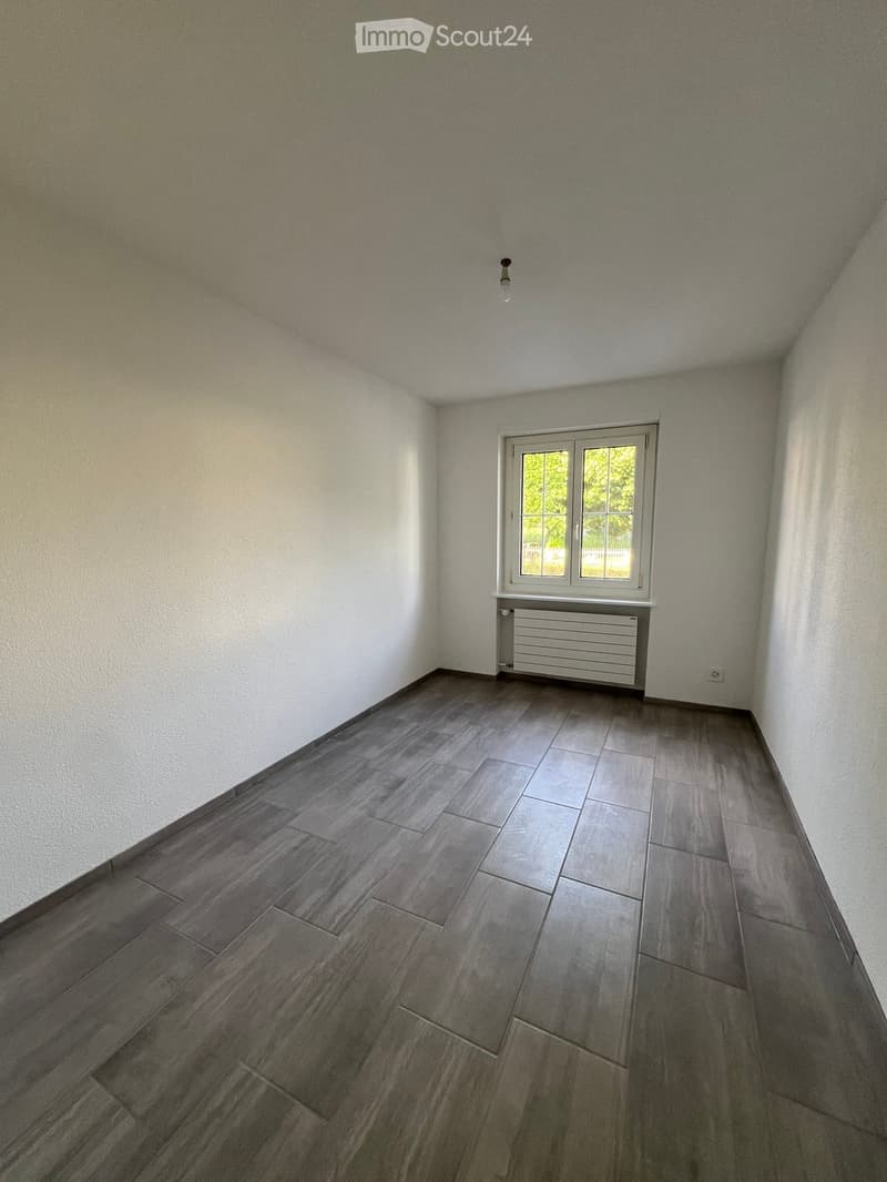 Empty room with light-colored walls, dark grey tiled floor, and a single window with white frame and radiator below it.