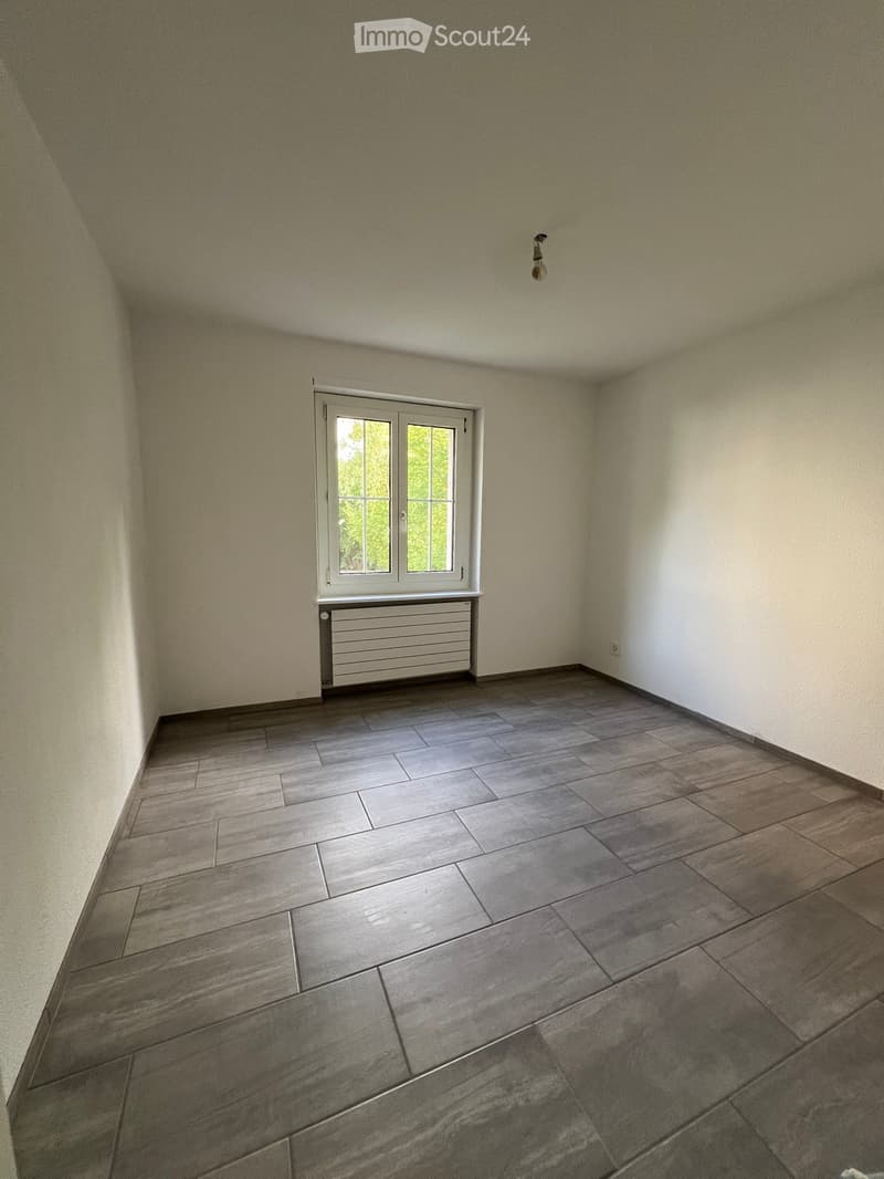 Empty room with grey tiled floor, white walls, a window with white frame, and a radiator below the window.