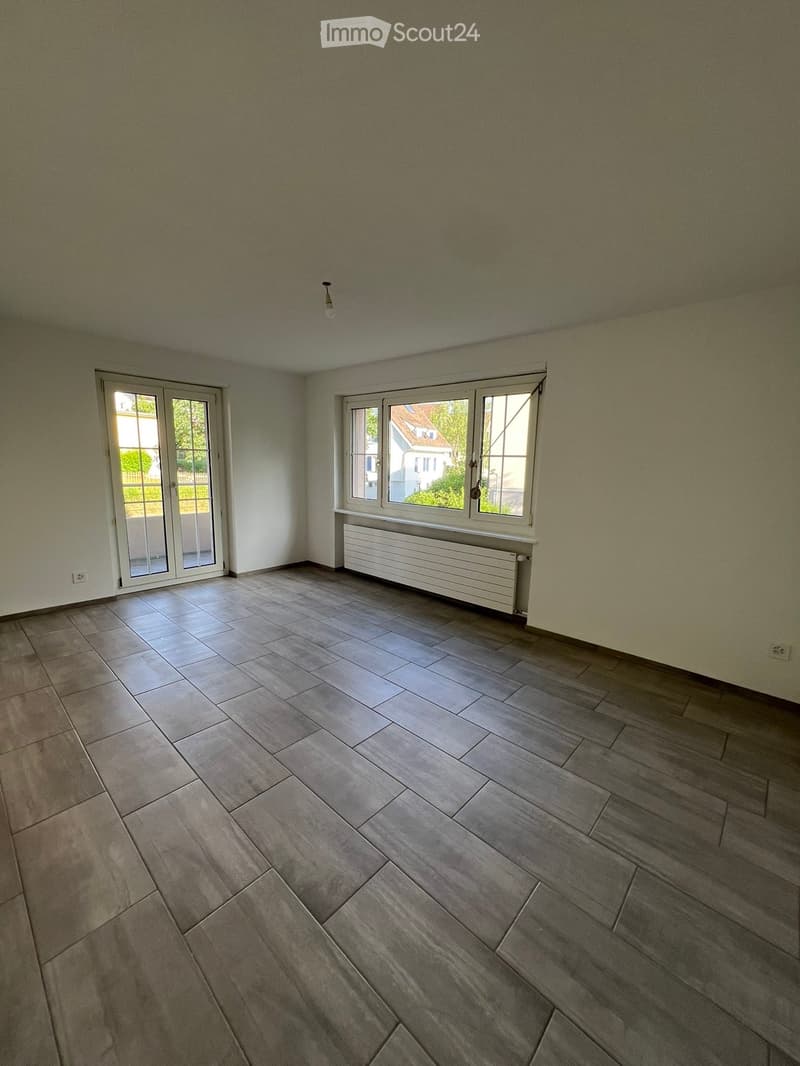 Empty room with light grey tiled flooring, white walls, and large windows with white frames. A radiator is visible beneath one window, and a ceiling light fixture is present.