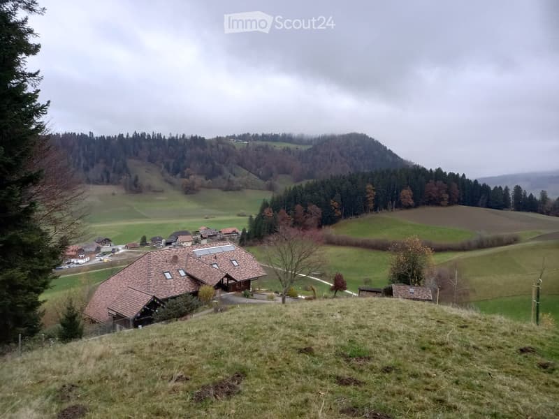 A two-story house with a red-tiled roof and solar panels, situated on a grassy hill with surrounding fields and trees.