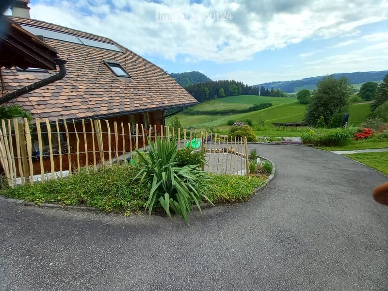 A modern house with a distinctive curved roof and brown tiled exterior, surrounded by a wooden fence and lush greenery, with a scenic rural landscape in the background.