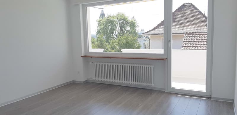Empty room with wooden flooring, white walls, large window, radiator, and sliding glass door leading to a balcony.