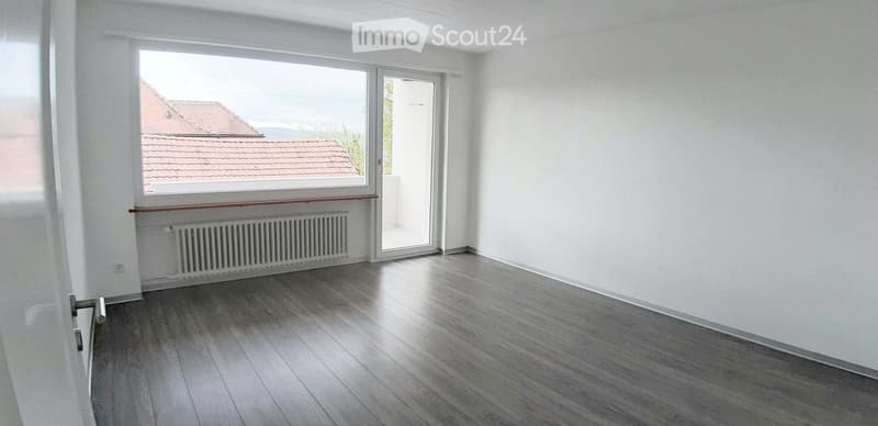 Empty room with wooden flooring, white walls, large window, radiator, and a door leading to a balcony.