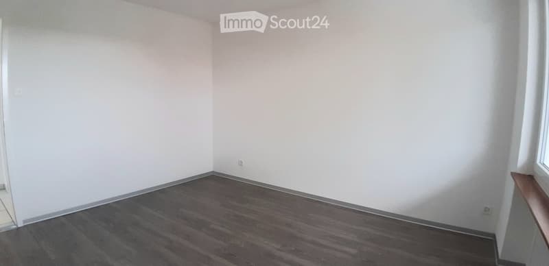 Empty room with white walls and dark wooden flooring, featuring a small window and a door frame visible.