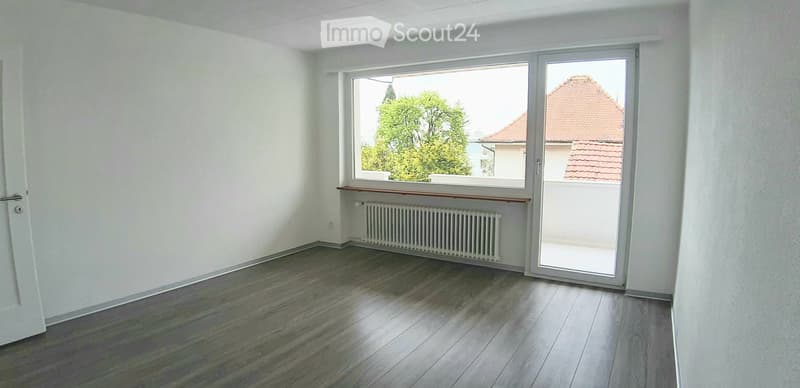 Empty room with wooden flooring, white walls, large window, and sliding glass door leading to a balcony.