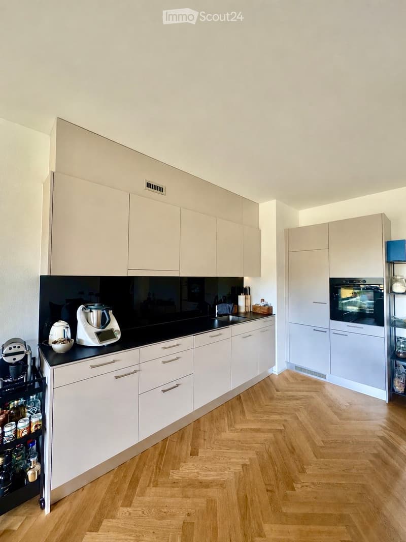 Modern kitchen with white cabinets, black countertop, wooden floor, and various appliances including a refrigerator, oven, blender, and coffee maker.
