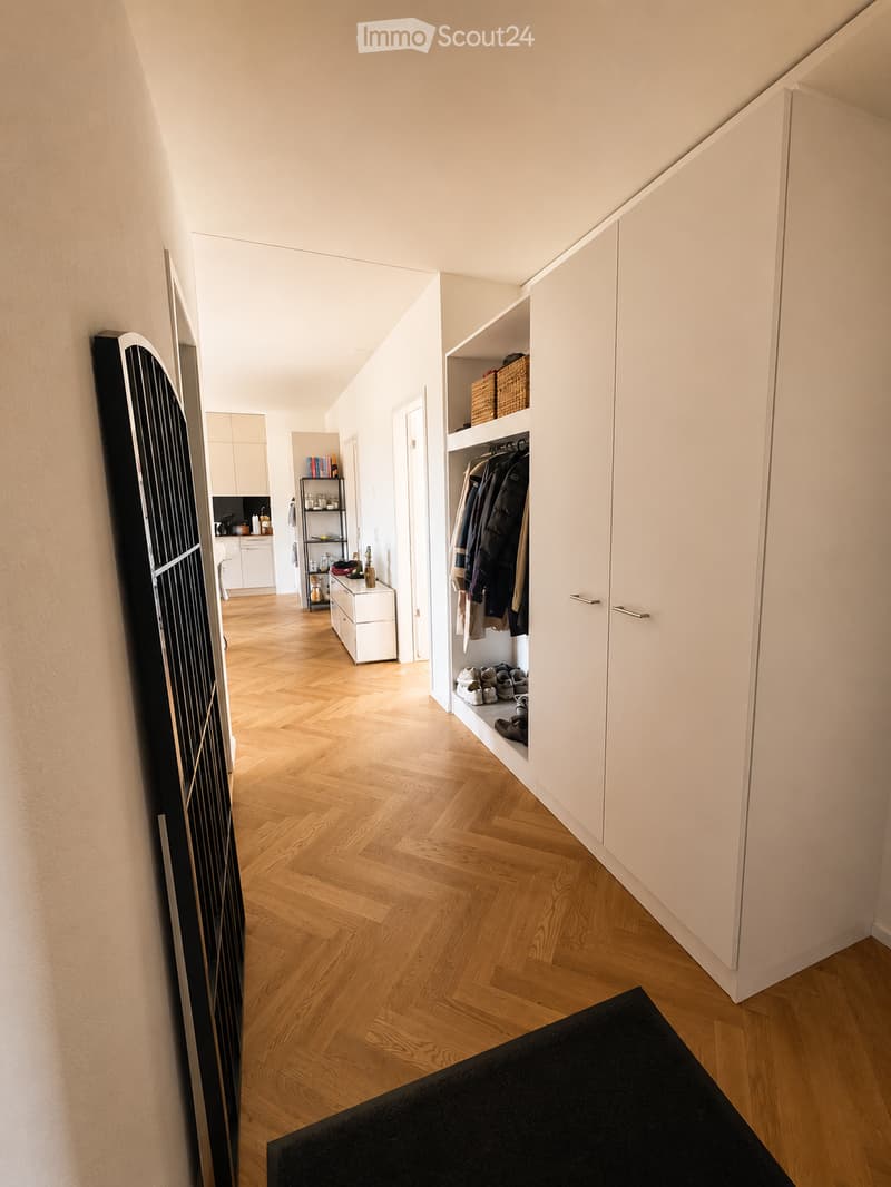 A modern hallway with wooden herringbone flooring, white walls, built-in closet with hanging space, and a small shelf with baskets. The hallway leads to a kitchen area with white cabinets and a countertop.