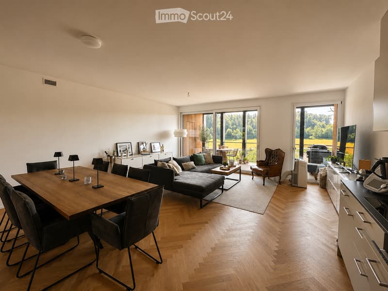 Open-plan living and dining area with wooden flooring, large windows, a sectional sofa, coffee table, dining table with chairs, and a lamp.