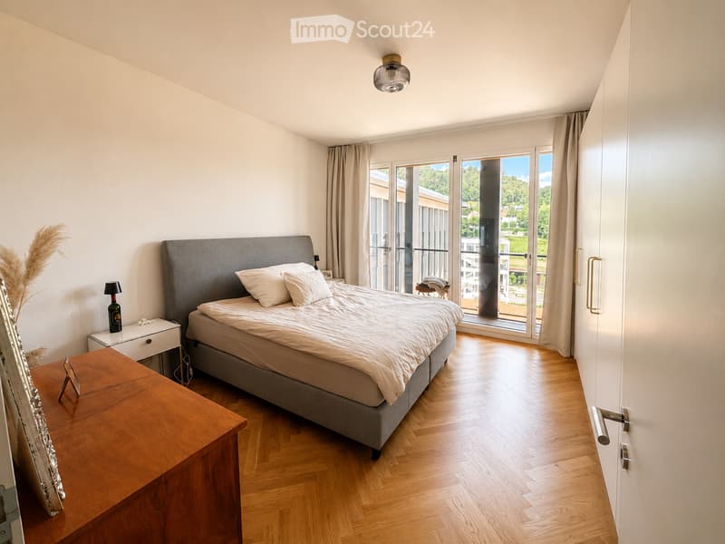 Modern bedroom with wooden flooring, large bed, nightstand, table, and view through large windows.