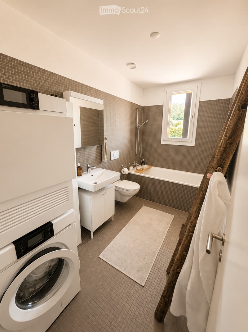 Modern bathroom with a white sink, toilet, bathtub, washing machine, and a large window letting in natural light. The space features tiled walls and a wooden towel rack with a towel.