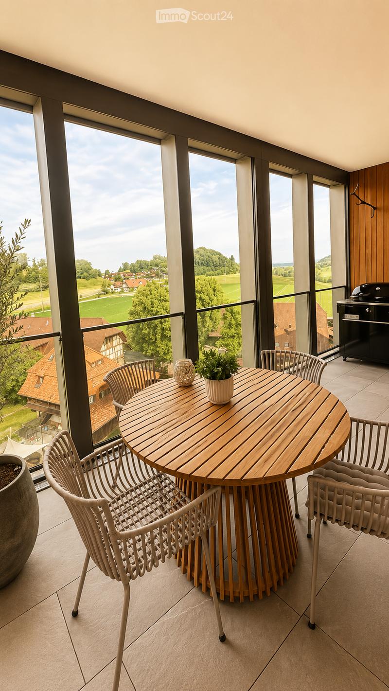 Balcony with a round wooden table, four white wicker chairs, a small potted plant, and a decorative vase. Large windows offer a scenic view of a countryside landscape.