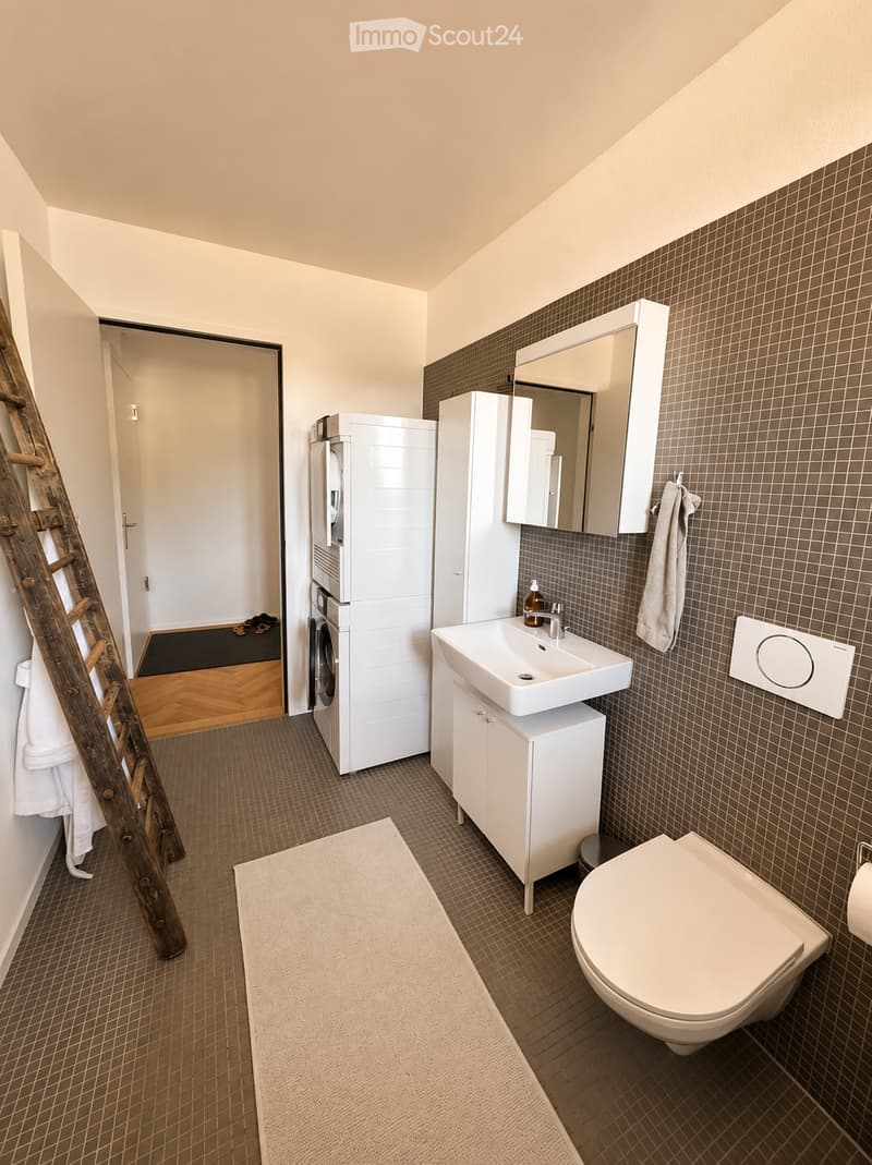 Modern bathroom with a white sink, toilet, mirror, and cabinet. Includes a washing machine in the adjacent space, a towel on a rack, and a bathmat on the floor.