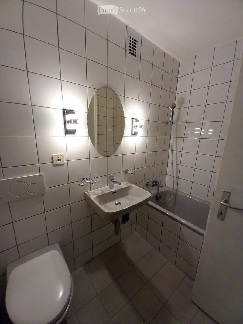 Modern bathroom with white tiled walls and floor, featuring a sink, toilet, bathtub, and showerhead. Includes a mirror and two cups on the sink counter.