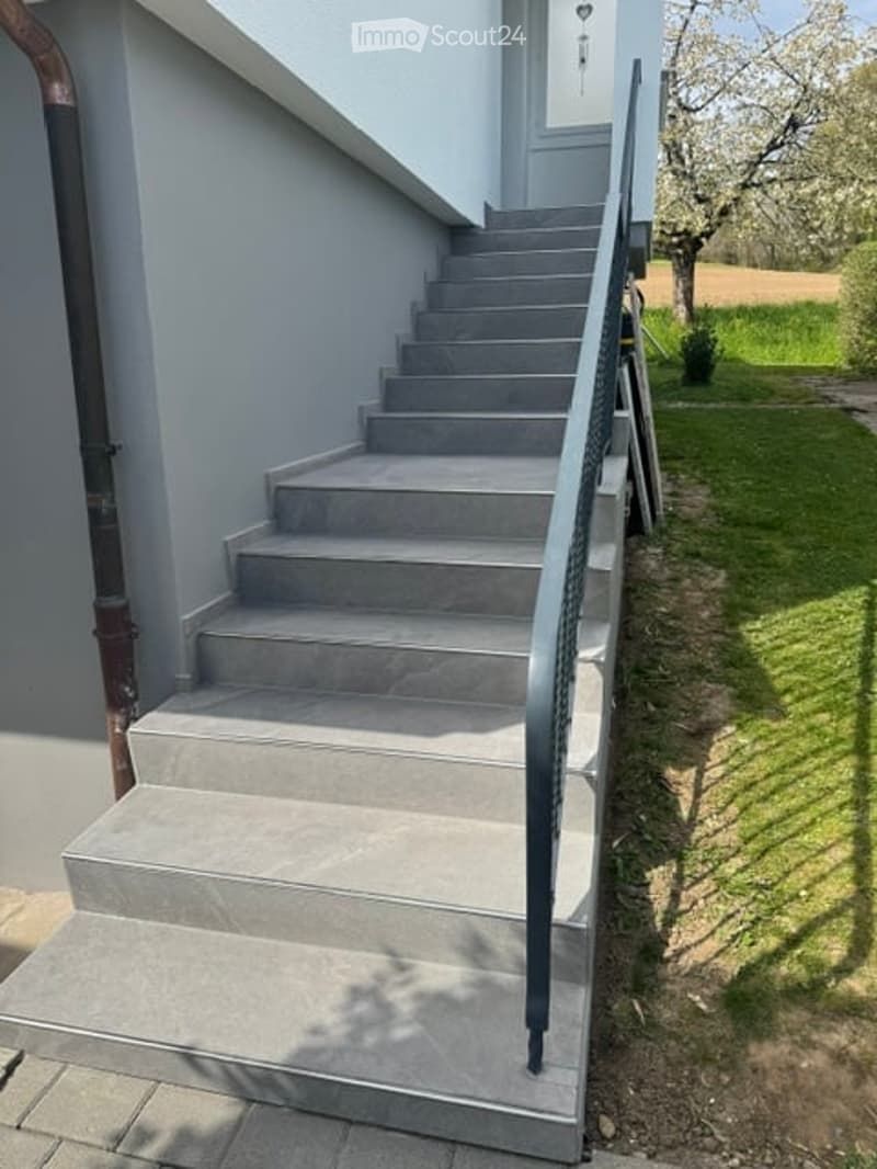 Concrete staircase leading up from a raised ground floor, with a metal handrail on one side and a grey wall on the other. The surrounding area includes a grassy lawn and a tree.