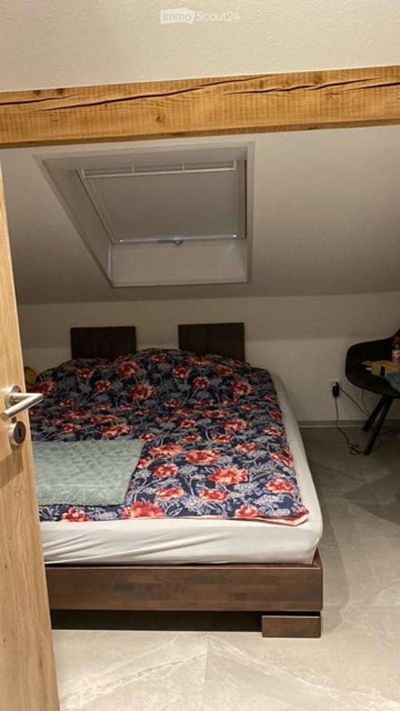 Cozy attic bedroom with a wooden bed, colorful floral bedding, a small nightstand, and a chair near a window with blinds.