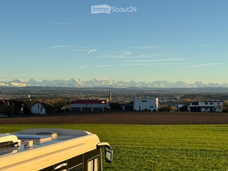 A wide, open field with a camper van in the foreground and a distant view of mountains and scattered houses.