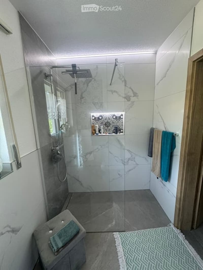 Modern bathroom with a large glass-enclosed shower, marble tiles, and a sleek design. Includes a showerhead, towel rack with towels, a small stool, and decorative bathroom accessories.