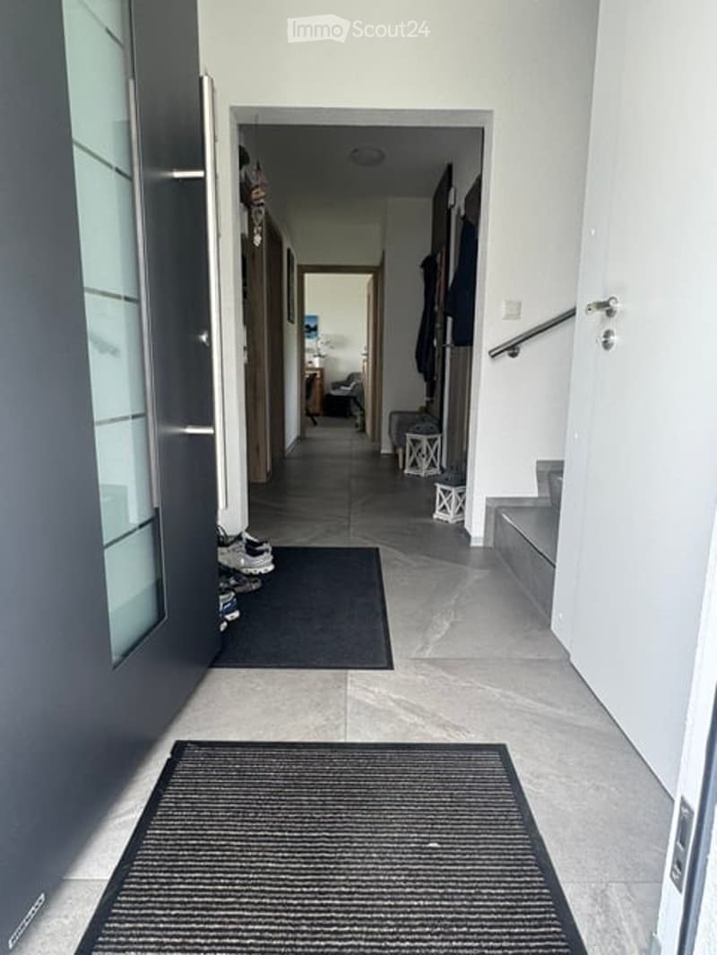 Modern hallway with tiled floor, white walls, and a staircase leading upwards. A small black doormat is placed at the entrance, and a coat rack with shoes is visible on the left side.
