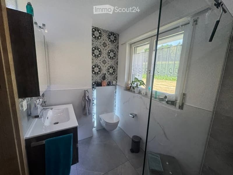 Modern bathroom with a white sink, mirror, toilet, and shower area. Features a decorative black and white patterned wall and a window with a view of the garden.