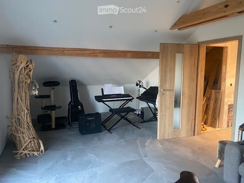 A cozy attic room with exposed wooden beams, a cat tree, a guitar, a keyboard on a stand, a speaker, and a chair. The room has large windows and a door leading to another area.