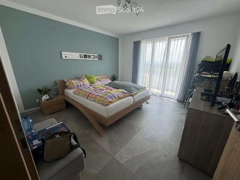 A modern bedroom with a double bed, nightstand, desk with a laptop, and a chair. The room features large windows with curtains, a grey wall, and a grey tiled floor.