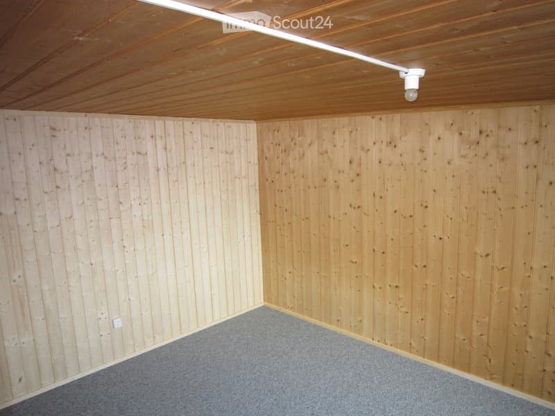 Empty room with wooden panel walls and a grey carpeted floor, featuring a wooden ceiling with a single light fixture.