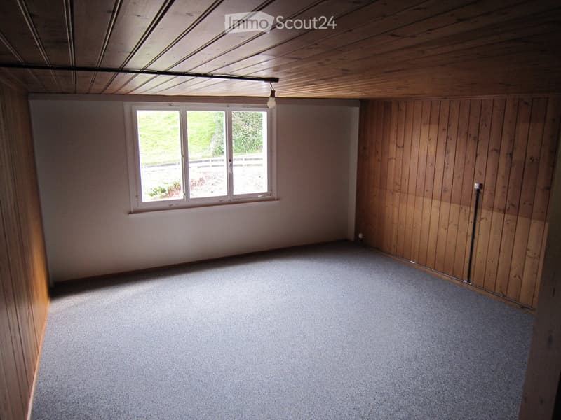 Empty room with wooden walls, carpeted floor, and a large window letting in natural light. The ceiling has exposed wooden beams and a single light fixture.