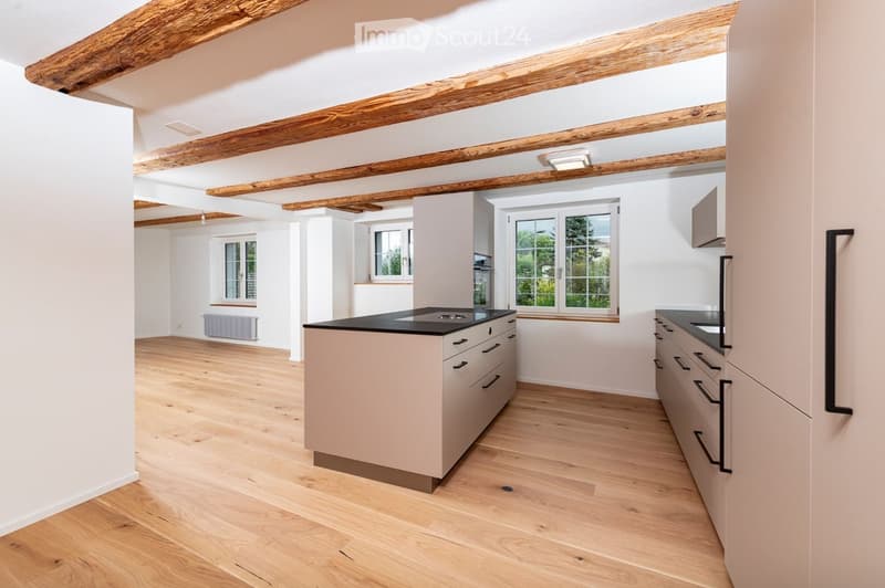 Bright, modern kitchen with light-colored cabinets, a central island, and wooden flooring. Features large windows and exposed wooden beams on the ceiling.