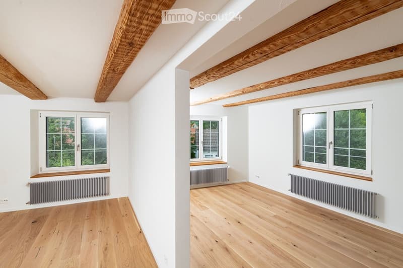 Bright, empty attic room with wooden floors, white walls, exposed wooden beams, and multiple windows offering natural light.