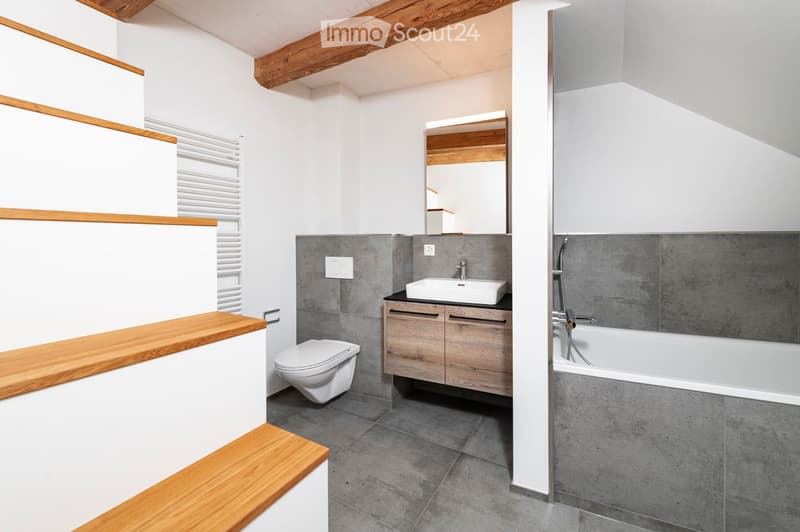 Modern bathroom with a toilet, sink, bathtub, and mirror. Features wooden beams on the ceiling and a wooden shelf on the left side.