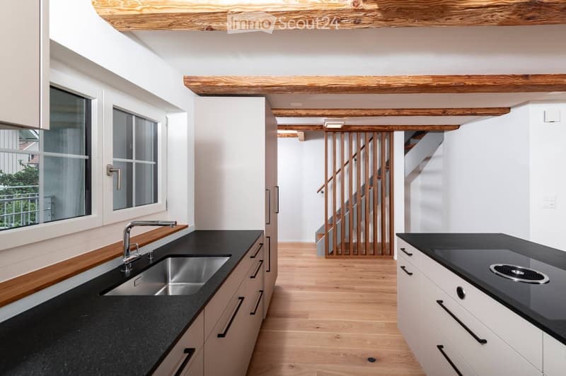 Modern kitchen with white cabinets, black countertops, stainless steel sink, and a visible staircase in the background.