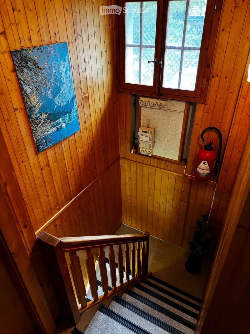 Wooden staircase with a handrail, carpeted steps, and a small window at the top. A painting and a fire extinguisher are visible on the walls.
