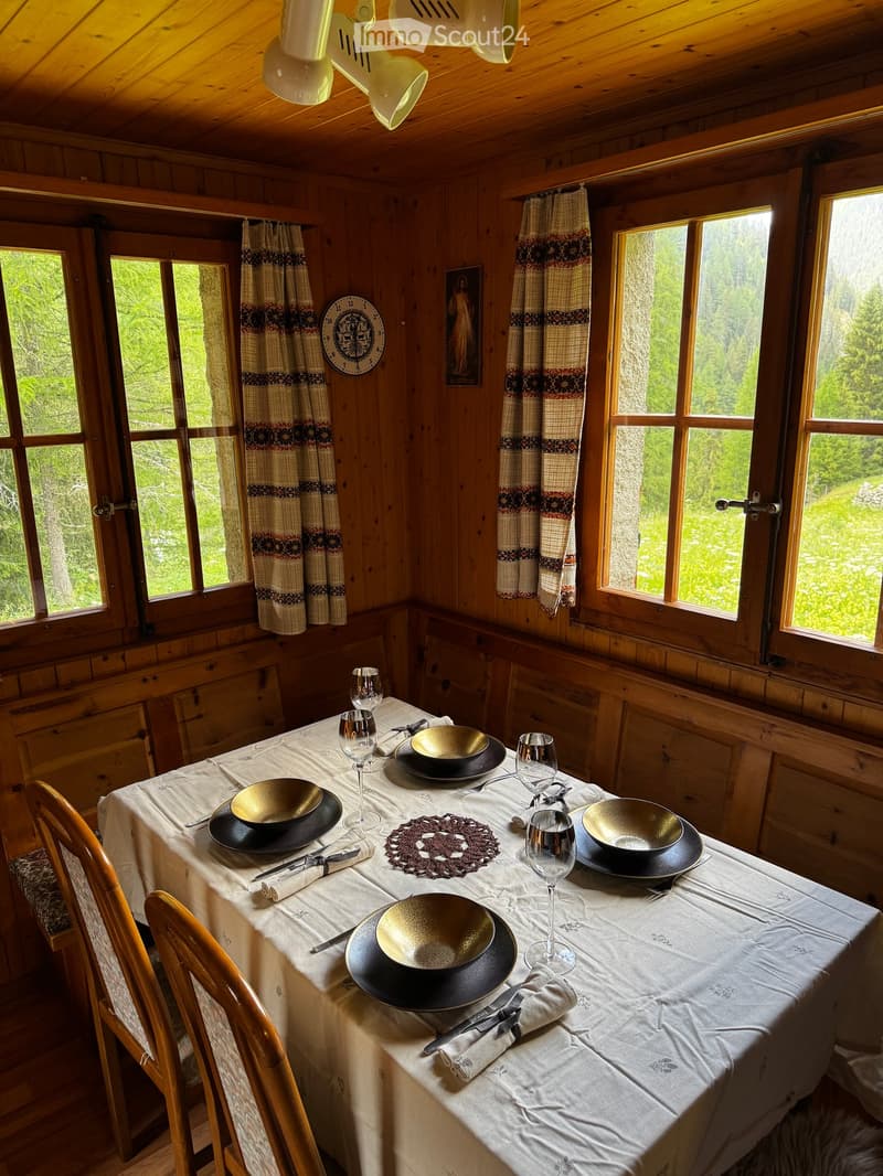 Cozy wooden kitchen with large windows, a dining table set for four with plates, bowls, glasses, and cutlery, and wooden chairs.