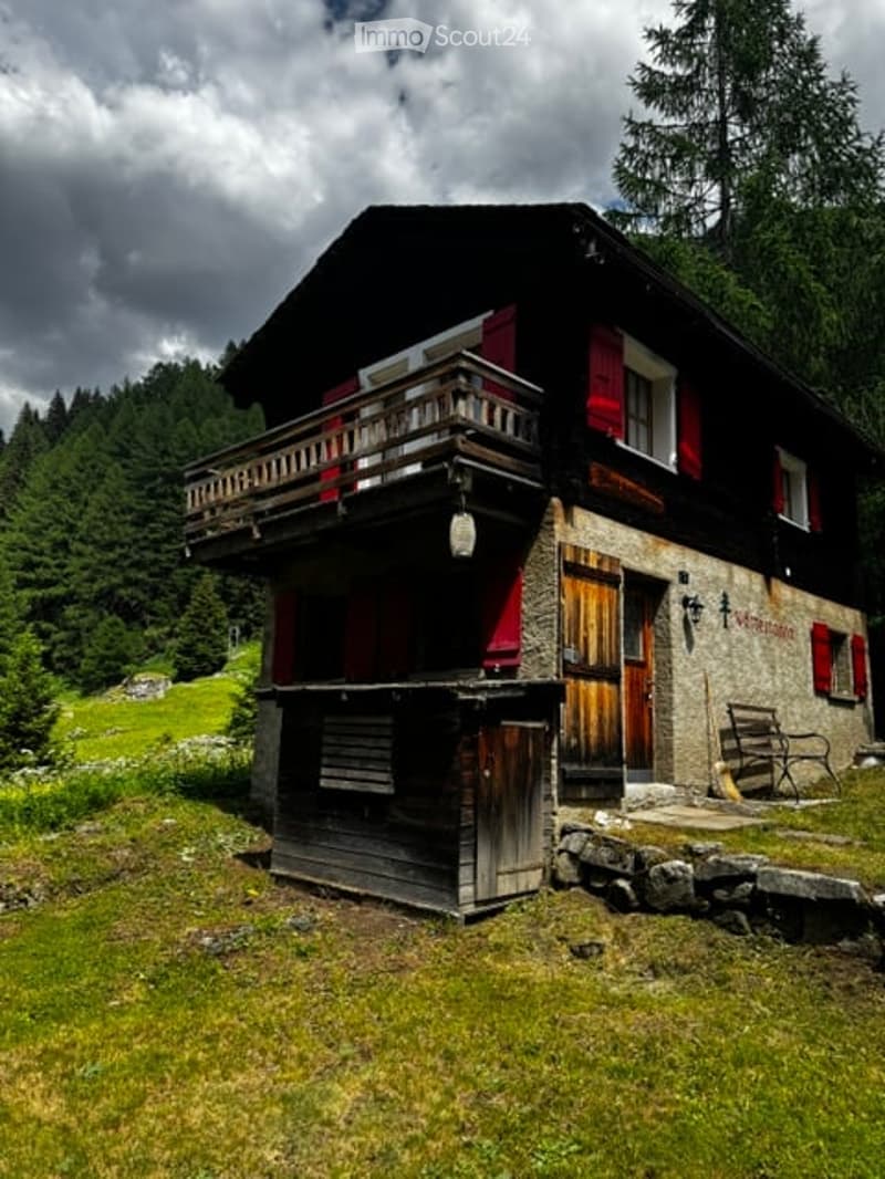 Traditional wooden chalet with red shutters, balcony, and stone base, surrounded by green forest and grassy area.