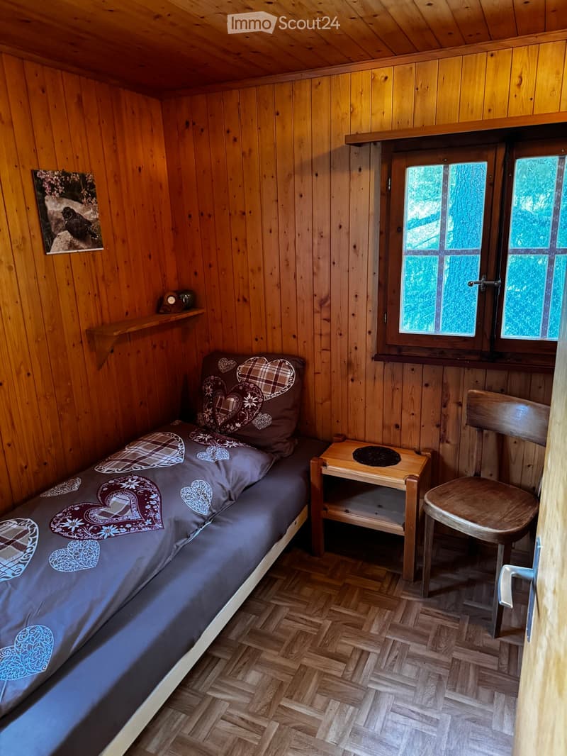 Cozy wooden bedroom with a single bed, nightstand, chair, and a small shelf with a clock and picture frame.