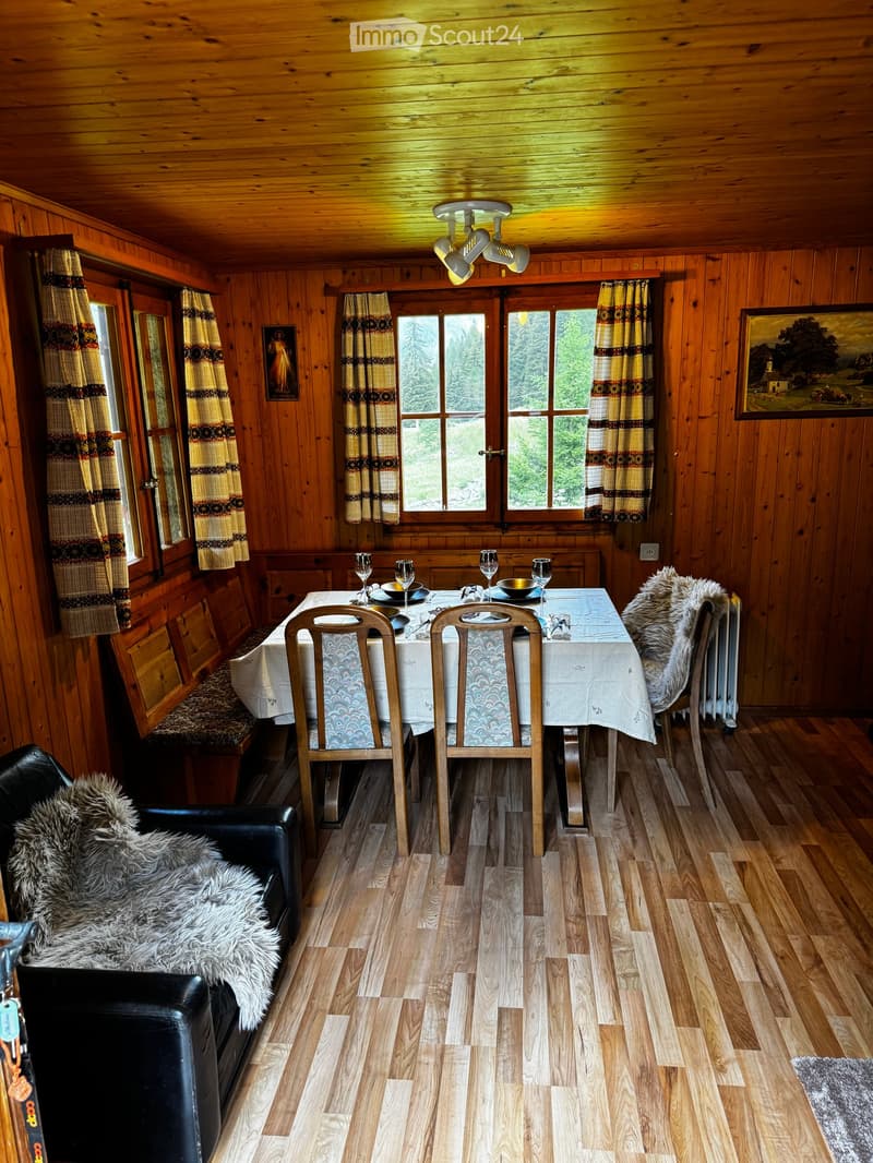 A cozy wooden-paneled room with a dining table, chairs, a sofa, and large windows with curtains.