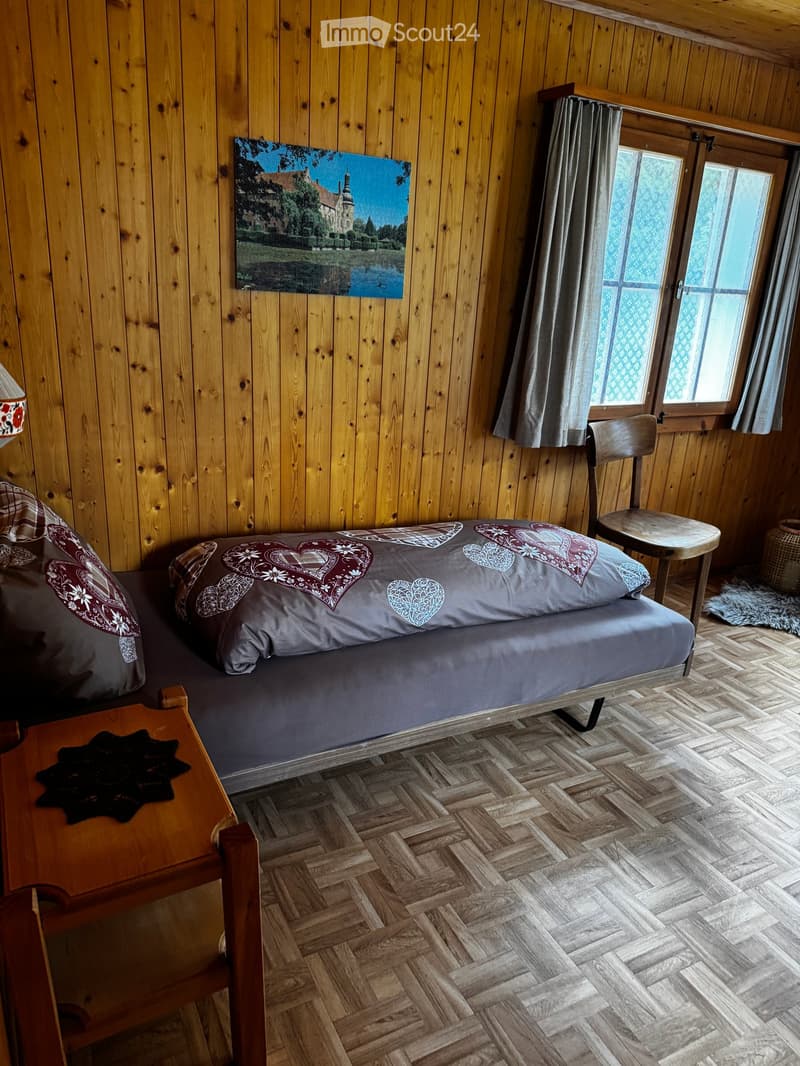 Cozy bedroom with wooden walls and herringbone wooden flooring, featuring a sofa bed with patterned cushions, a small wooden table, and a picture frame on the wall.
