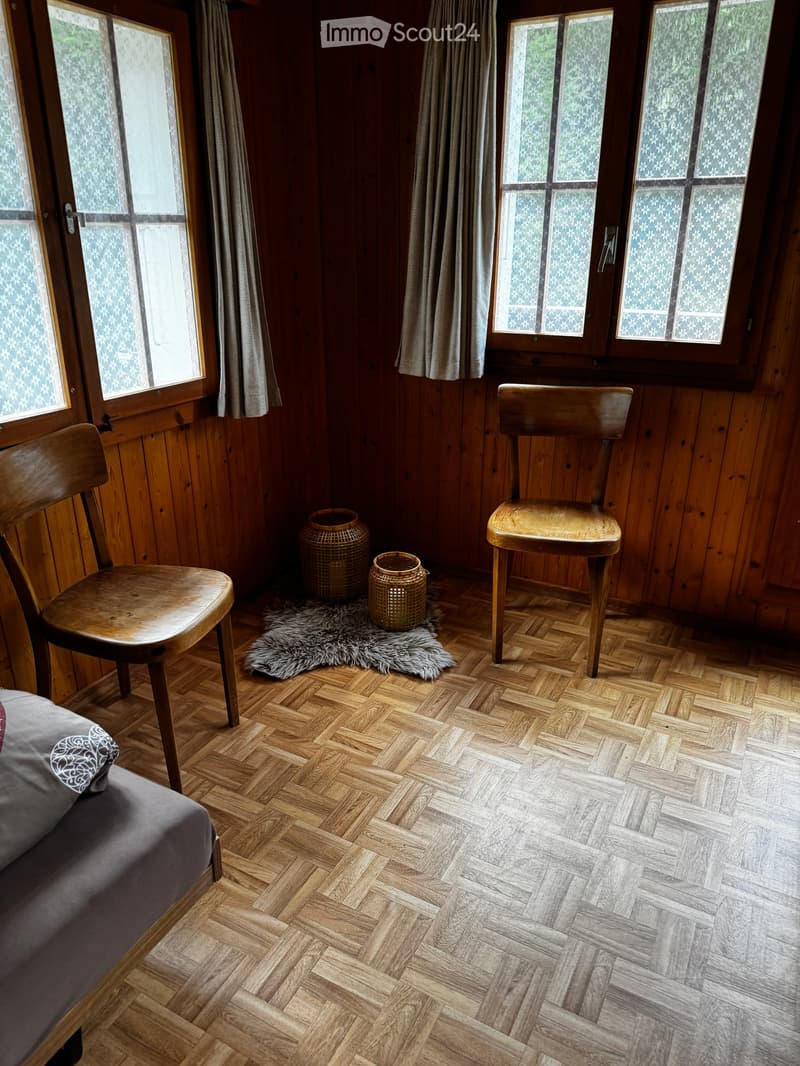 Cozy bedroom with wooden herringbone flooring, large windows with curtains, two wooden chairs, a small rug with two vases, and a bed with a grey pillow.
