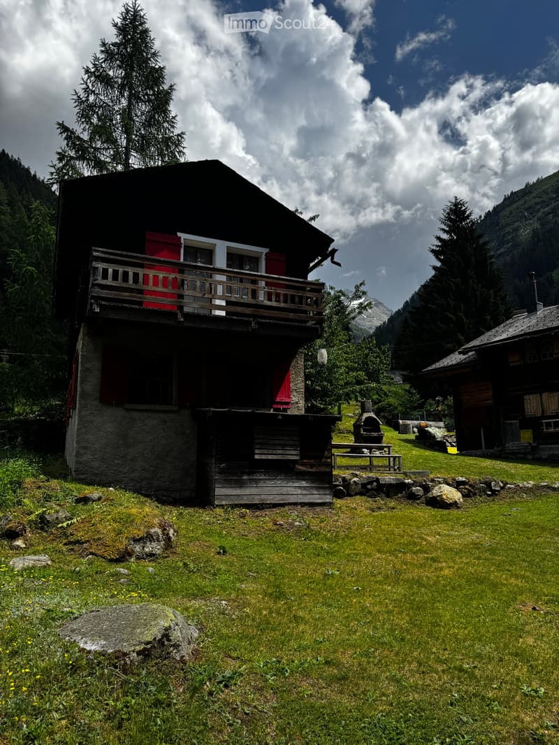 A two-story wooden house with a balcony, red shutters, and a stone base, situated in a grassy area surrounded by trees and mountains.