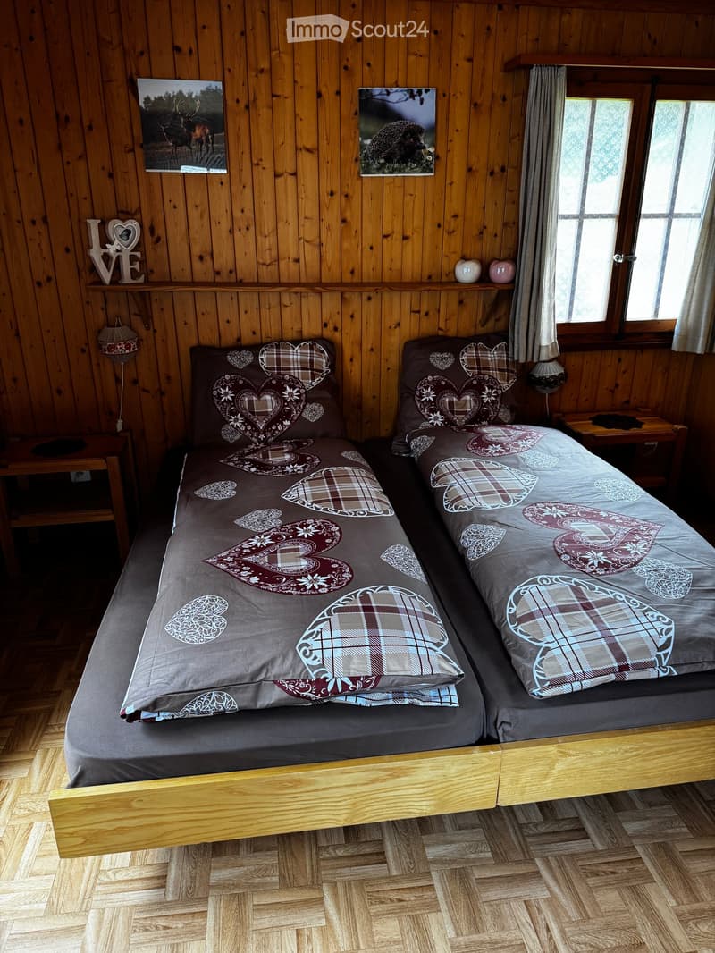 Cozy bedroom with wooden walls and floors, featuring a double bed with patterned bedding, two nightstands with lamps, and decorative picture frames on the wall.