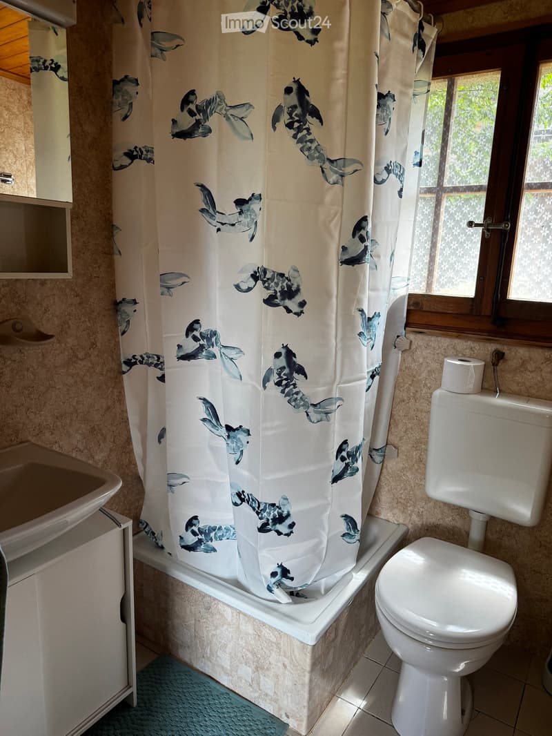 Bathroom with a bathtub, toilet, sink, and a shower curtain featuring fish designs. The room has tiled walls and a window with wooden framing.
