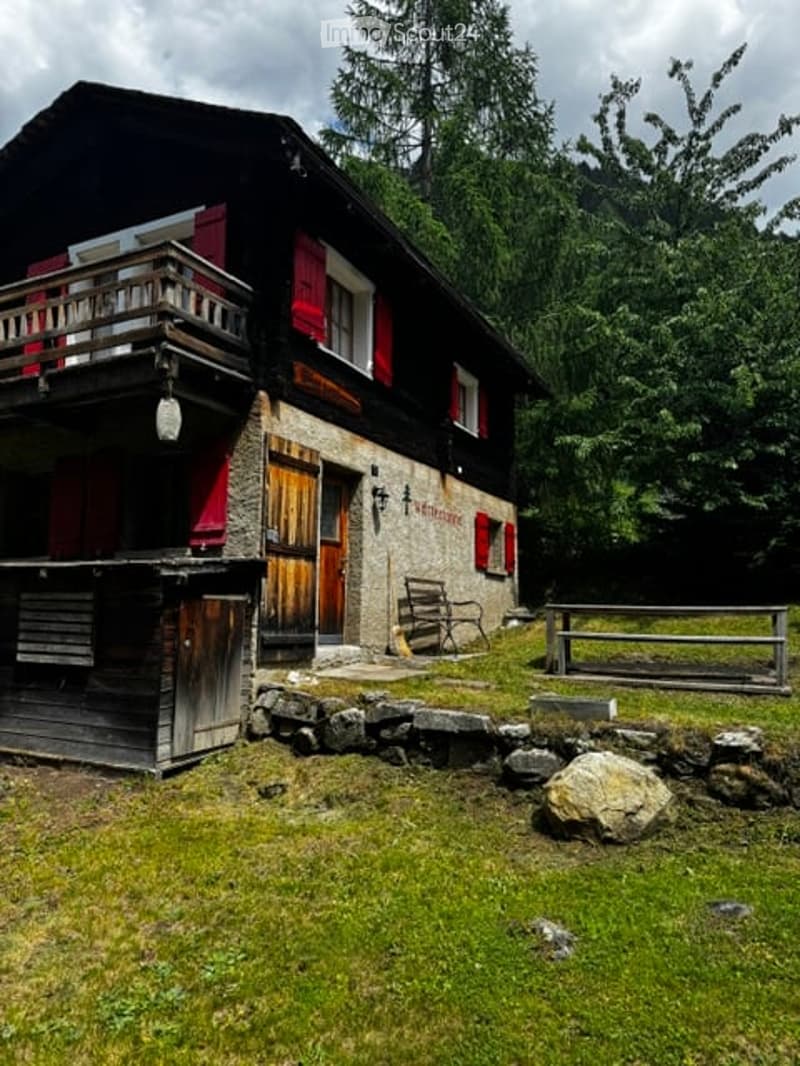 A rustic wooden house with red shutters, a balcony, and a small outdoor seating area. The house is surrounded by greenery and features a stone pathway leading to the entrance.