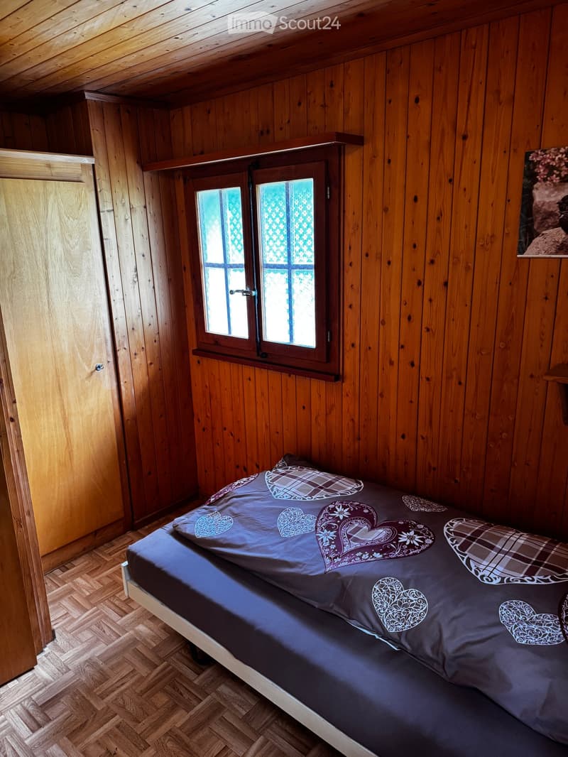 Cozy bedroom with wooden walls and slanted ceiling, featuring a single bed with patterned bedding and a small window with decorative glass panes.