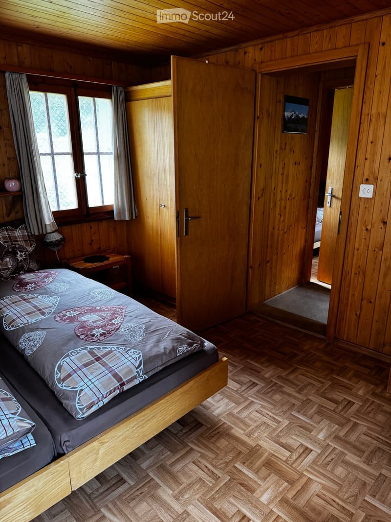 Cozy wooden bedroom with a double bed, nightstand, and large windows with curtains. The room features wooden walls and a herringbone parquet floor.